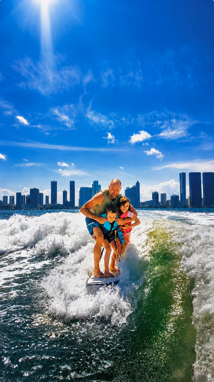 Un papá haciendo wakesurf con sus dos hijos agarrados a él, el skyline de Miami al fondo